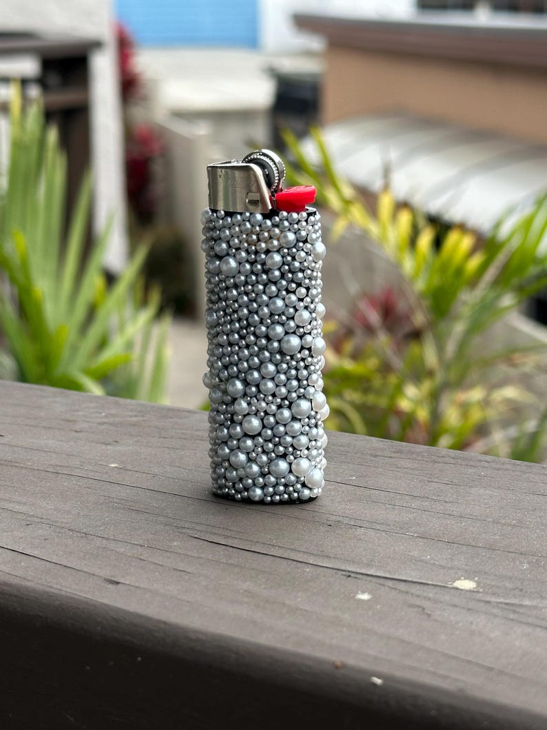 Puede incluir: Un encendedor plateado cubierto con perlas de imitaci&oacute;n de varios tama&ntilde;os. La parte superior del encendedor tiene una carcasa de metal plateado con un bot&oacute;n de encendido rojo. El encendedor est&aacute; de pie sobre una superficie de madera.