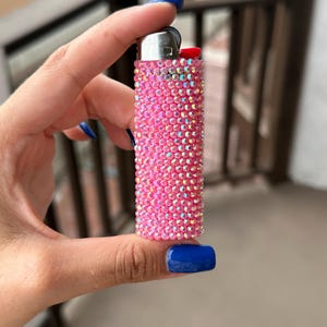 Puede incluir: Un encendedor rosa completamente incrustado con pedrer&iacute;a iridiscente. El encendedor tiene una parte superior de metal plateado y un bot&oacute;n rojo. El encendedor se sostiene en una mano con esmalte de u&ntilde;as azul.