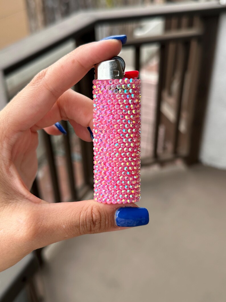 Puede incluir: Un encendedor rosa completamente incrustado con pedrer&iacute;a iridiscente. El encendedor tiene una parte superior de metal plateado y un bot&oacute;n rojo. El encendedor se sostiene en una mano con esmalte de u&ntilde;as azul.