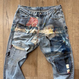 Könnte beinhalten: Hellblaue Jeans mit mehreren Patches und Farbspritzern. Zu den Patches gehören ein rosa Blumengewebe und verschiedene Jeansstücke. Die Jeans haben einen Used-Look mit entspannter Passform und umgeschlagenen Säumen.