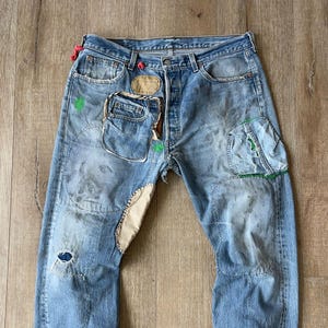 Könnte beinhalten: Hellblaue Jeans mit mehreren Flicken und Reparaturen. Die Jeans haben einen Used-Look mit sichtbaren Gebrauchsspuren, darunter Flicken aus beigem Stoff und grüne Farbakzente. Die Taschen sind ebenfalls geflickt.