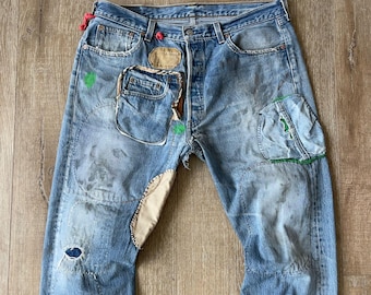 May include: Light blue denim jeans with multiple patches and repairs. The jeans feature a distressed look with visible wear and tear, including patches of beige fabric and green paint accents. The pockets are also patched.