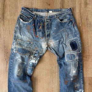 Könnte beinhalten: Verblasste blaue Jeans mit einem Used-Look, mit mehreren Patches und sichtbaren Nähten. Die Jeans haben weiße Farbspritzer und einen Knopfverschluss. Auf Knie und Oberschenkel sind Patches aufgenäht.
