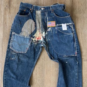 May include: Distressed blue denim jeans with multiple patches and stitching details. The jeans have a faded look and are decorated with a small American flag patch.