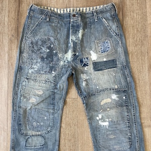 May include: Distressed blue denim jeans with multiple patches and white paint splatters. The jeans have a relaxed fit and a straight leg.