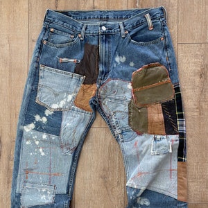 Peut inclure: Un jean en denim bleu avec de multiples patchs de tissus et de couleurs différents, dont du cuir marron, de la toile verte et du plaid. Le jean a un aspect vieilli avec des zones délavées et des éclaboussures de peinture blanche.