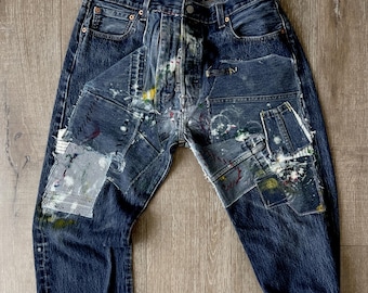 May include: Distressed blue denim jeans with multiple patches and paint splatters. The jeans have a relaxed fit and a cuffed hem.