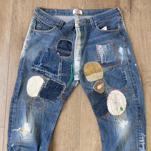 Op de afbeelding: Blauwe denim jeans met meerdere patches die erop zijn genaaid. De patches zijn gemaakt van verschillende stoffen en kleuren, waaronder bruin, wit en groen. De jeans hebben een verweerde look met scheuren en gaten.