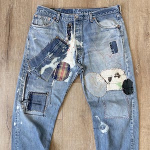 May include: A pair of blue denim jeans with multiple patches sewn on. The patches are made of different fabrics, including plaid, denim, and canvas. The jeans have a faded and distressed look, with white paint splatters throughout.