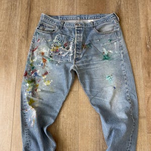 May include: Blue denim jeans with paint splatters in various colours, including red, yellow, green, and blue. The jeans have a single button closure and a five-pocket design.