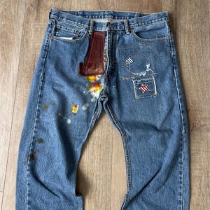 Op de afbeelding: Blauwe denim jeans met verfspatten en patches. De patches omvatten een rood, wit en blauwe ster, een zwart-witte cirkel en een rood-wit gestreepte patch.
