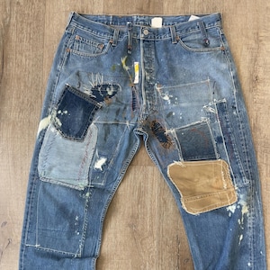 Op de afbeelding: Blauwe denim jeans met meerdere patches en verfspatten. De patches zijn gemaakt van verschillende stoffen en kleuren, waaronder bruin, blauw en wit. De jeans heeft een ontspannen pasvorm en een rechte pijp.