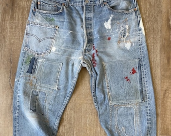 May include: Light blue denim jeans with multiple patches and paint splatters. The jeans have a faded look and are distressed.