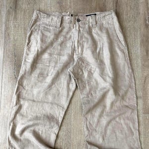 May include: Light beige wide-leg trousers with several patch pockets. The trousers feature a button and zip fastening, along with belt loops. The material looks like a light linen or cotton mix, ideal for casual wear.