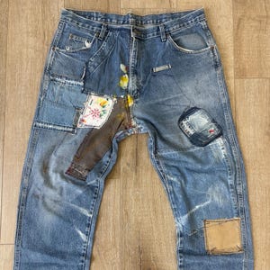 Könnte beinhalten: Blaue Jeans aus Denim mit mehreren aufgenähten Patches. Die Patches bestehen aus verschiedenen Stoffen und Farben, darunter braunes Leder, blauer Denim und ein Blumendruck.