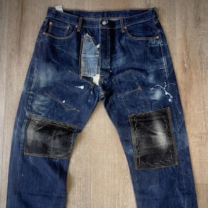 Puede incluir: Vaqueros de mezclilla azul desgastados con parches de cuero negro en las rodillas. Los vaqueros tienen un aspecto descolorido y desgastado, con costuras y reparaciones visibles.