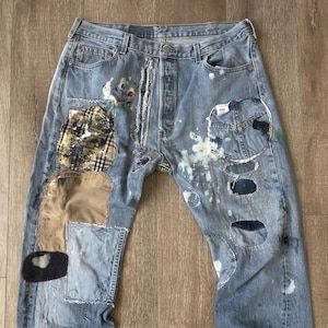 May include: Light blue denim jeans with various patches and paint splatters. Patches feature plaid, tan, and blue fabrics. The jeans have a button fly and distressed details, presenting a unique, artistic design.