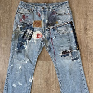 Peut inclure: Un jean en denim bleu clair avec plusieurs patchs et des éclaboussures de peinture. Le jean a une fermeture éclair sur la jambe droite et un patch en cuir marron avec le texte "Levi's" sur le devant.