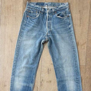 May include: Light blue denim jeans with a button fly and classic five-pocket design. The jeans show signs of wear and fading, with a straight leg cut. The jeans are laid flat on a wooden surface.