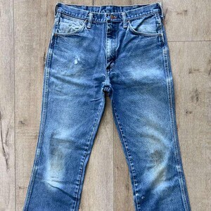 May include: Vintage blue denim jeans with a classic five-pocket design. The jeans show signs of wear and fading, with lighter patches on the thighs and knees. The jeans have a straight leg cut and a button and zip closure.