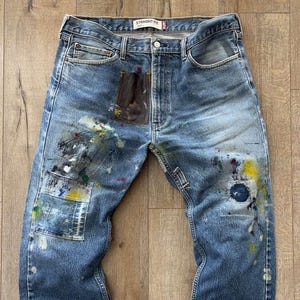 Puede incluir: Vaqueros azules de mezclilla con salpicaduras de pintura blanca, amarilla y verde. Los vaqueros tienen un corte recto y un parche marrón en la parte delantera. El texto "STRAIGHT FIT" es visible.