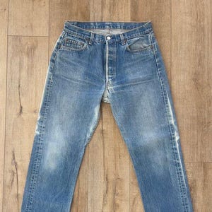 May include: Faded blue denim jeans with a button fly. The jeans have a classic five-pocket design and show signs of wear, with lighter areas on the thighs and knees. The jeans are laid flat on a wooden surface.