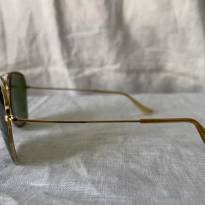 May include: Gold-framed aviator sunglasses with green lenses. The sunglasses have a classic design with a double bridge and a teardrop shape. The temples are thin and gold-colored, with a curved earpiece.