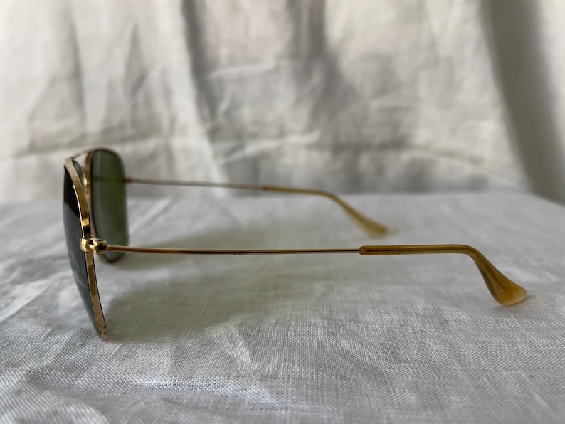May include: Gold-framed aviator sunglasses with green lenses. The sunglasses have a classic design with a double bridge and a teardrop shape. The temples are thin and gold-colored, with a curved earpiece.