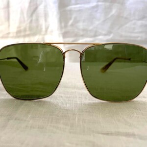 May include: Gold-framed aviator sunglasses with green lenses. The sunglasses have a double bridge and a classic design. The lenses are a solid green color, and the frames are a shiny gold. The sunglasses are displayed on a white surface.