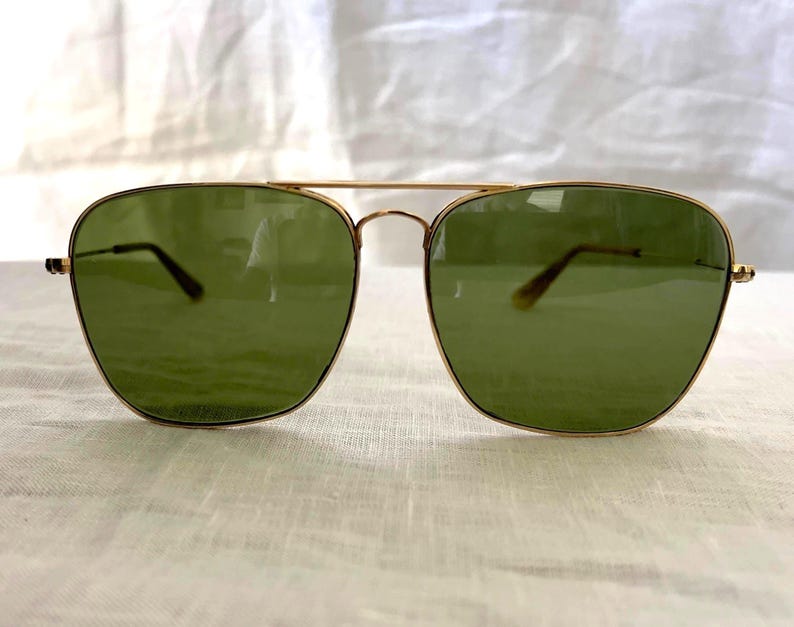 May include: Gold-framed aviator sunglasses with green lenses. The sunglasses have a double bridge and a classic design. The lenses are a solid green color, and the frames are a shiny gold. The sunglasses are displayed on a white surface.