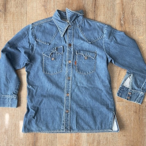 May include: A light blue denim shirt with long sleeves, two chest pockets, and a button-down front. The shirt has a pointed collar and decorative stitching. A small orange tag is visible on the left pocket.