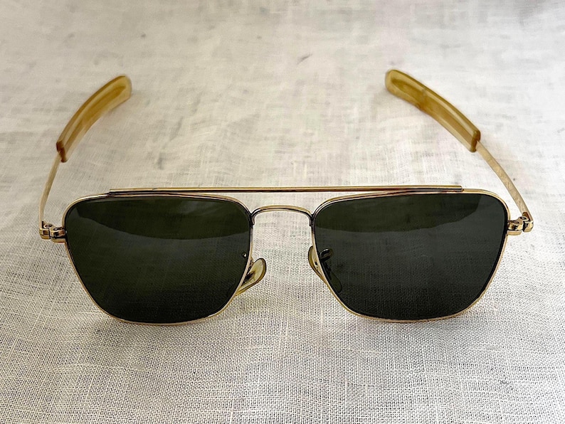 Puede incluir: Gafas de sol estilo aviador con montura de metal dorado y lentes verde oscuro. Las patillas son de color marr&oacute;n claro. Las gafas se muestran sobre una superficie texturizada blanquecina.