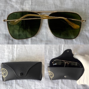 May include: Gold-framed sunglasses with green lenses, displayed with a black leather case. The sunglasses have a double bridge and a classic aviator style. The case features a button closure and a gold logo.