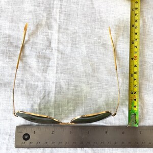 May include: Gold-framed sunglasses with green lenses are displayed. The sunglasses are measured with a ruler, showing a length of approximately 6 inches. The glasses have a classic aviator style.