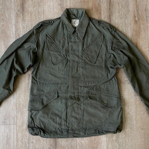 May include: An olive green military-style jacket with a button-down front, two chest pockets, and two lower pockets. The jacket has a collar and long sleeves. The garment is laid flat on a wooden surface.