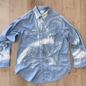 Könnte beinhalten: Ein langärmeliges hellblaues Jeanshemd mit einem gebleichten Muster aus unregelmäßigen weißen Flecken auf dem Stoff. Das Hemd verfügt über eine durchgehende Knopfleiste, einen spitzen Kragen, zwei Brusttaschen mit Klappen und geknöpfte Manschetten.