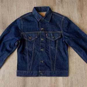 以下が含まれることがあります： ボタン留めの濃い青色のデニムジャケット。胸ポケットが2つ、小さな赤いタグが付いています。ジャケットはクラシックな襟で、丈夫なデニム素材で作られています。木製の表面に平らに置かれています。