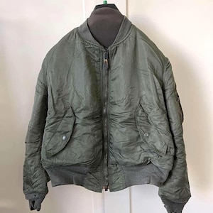 May include: An olive green bomber jacket with a full zipper closure, two front pockets, and a ribbed collar, cuffs, and hem. The jacket is hanging on a metal hanger against a white background. The jacket appears to be made of a durable, possibly nylon, material.