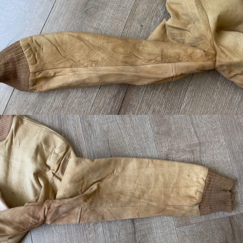 May include: Close-up of a tan leather jacket sleeve. The sleeve features a ribbed brown cuff and a weathered appearance. The jacket appears to be vintage or distressed, with visible wear and tear. The leather has a warm, golden hue.