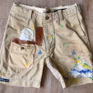 May include: Khaki-colored shorts with paint splatters in various colors. The shorts have a front pocket with a button closure and a leather patch. The shorts are distressed and show signs of wear.