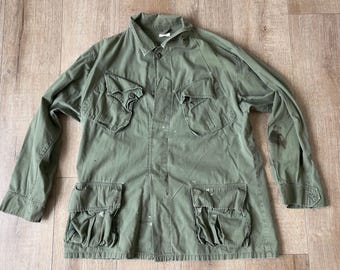 Könnte beinhalten: Olivgrüne Jacke im Militärstil mit vier Vordertaschen und einem Knopfverschluss. Die langärmlige Jacke hat einen Kragen und scheint aus einem strapazierfähigen Stoff zu sein. Einige Farbflecken sind auf der Jacke sichtbar.
