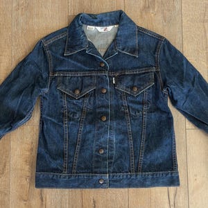 May include: A dark blue denim jacket with a button-up front, two chest pockets, and two side pockets. The jacket has a classic collar and long sleeves with buttoned cuffs. The Levi's brand label is visible on the jacket.