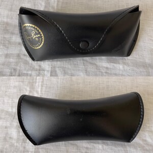 Może przedstawiać: Czarne etui na okulary przeciwsłoneczne Ray-Ban, zamknięte i otwarte. Zamknięte etui ma zapięcie na zatrzask i złoty emblemat z logo Ray-Ban. Otwarte etui ma zakrzywiony kształt.