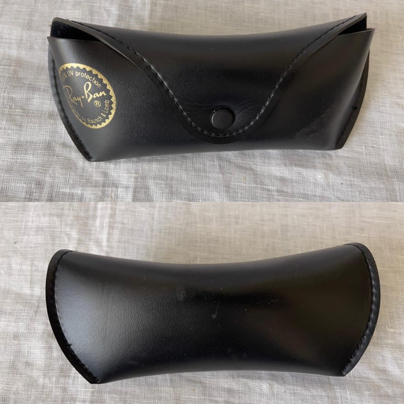 Może przedstawiać: Czarne etui na okulary przeciwsłoneczne Ray-Ban, zamknięte i otwarte. Zamknięte etui ma zapięcie na zatrzask i złoty emblemat z logo Ray-Ban. Otwarte etui ma zakrzywiony kształt.