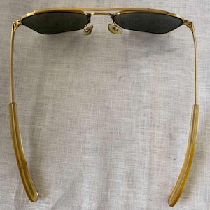 Puede incluir: Gafas de sol con montura dorada y lentes oscuras. Las gafas de sol tienen un estilo aviador cl&aacute;sico con doble puente y patillas curvas. Las lentes son de color verde oscuro y la montura es dorada brillante.