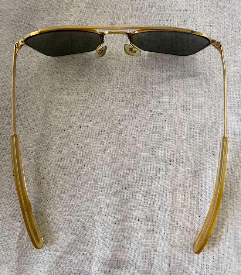 Puede incluir: Gafas de sol con montura dorada y lentes oscuras. Las gafas de sol tienen un estilo aviador cl&aacute;sico con doble puente y patillas curvas. Las lentes son de color verde oscuro y la montura es dorada brillante.