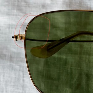 May include: Close-up of aviator sunglasses with gold-colored frames and green lenses. The glasses have a classic design, with a double bridge and a thin, curved temple. The lenses are slightly tinted, providing a vintage aesthetic.