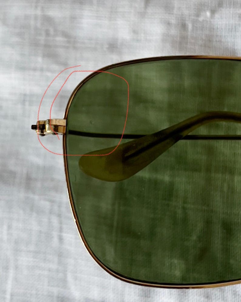 May include: Close-up of aviator sunglasses with gold-colored frames and green lenses. The glasses have a classic design, with a double bridge and a thin, curved temple. The lenses are slightly tinted, providing a vintage aesthetic.