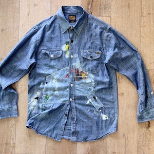 Könnte beinhalten: Ein blaues langärmeliges Hemd mit Knopfleiste und Used-Look, verziert mit Farbspritzern in Weiß, Gelb und Grün. Das Hemd verfügt über zwei Brusttaschen, einen spitzen Kragen und ein zentrales Aufnäher-Detail mit einem kleinen Grafikdruck.