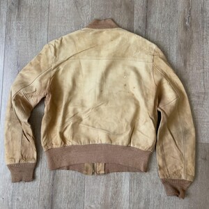 May include: A tan leather bomber jacket with a brown knit collar, cuffs, and waistband. The jacket has a zipper closure and visible wear and tear, giving it a vintage look. The jacket is laid flat on a wooden surface.
